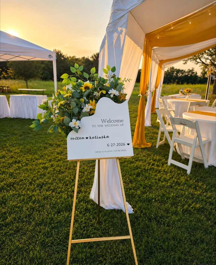 Wedding welcome sign with sunflowers beside a draped tent at sunset